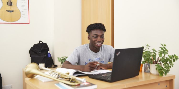 A boy is sat at a desk, smiling at his laptop